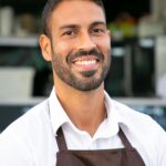 Smiling male café worker in apron, standing confidently outdoors in a business setting.